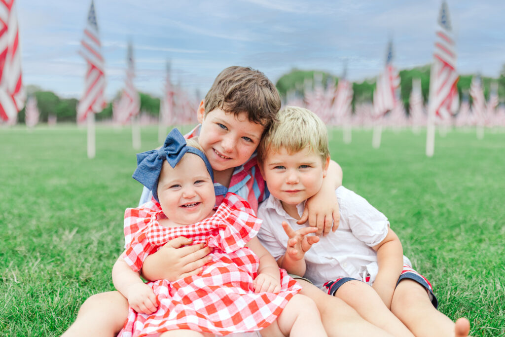 A joyful moment captured by Bridgette Brown Photography at Seven Gables Park in Wheaton, Illinois. Two children, dressed in festive red, white, and blue attire, are smiling and playing near the Freedom Flag Display. The vibrant American flags and lush greenery create a picturesque and patriotic backdrop, embodying the spirit of the Fourth of July. The image exudes family love, patriotism, and the beauty of the holiday celebration.